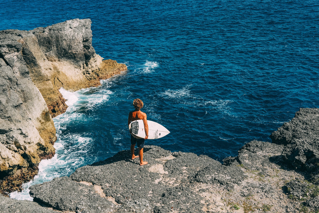 Surfer holding surfboard, gazing at the ocean while standing on a rocky cliff in the sunlight.