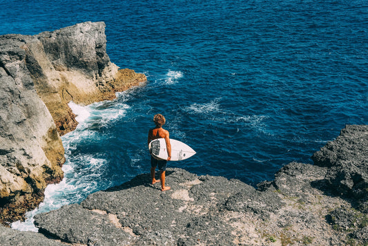 Surfer holding surfboard, gazing at the ocean while standing on a rocky cliff in the sunlight.