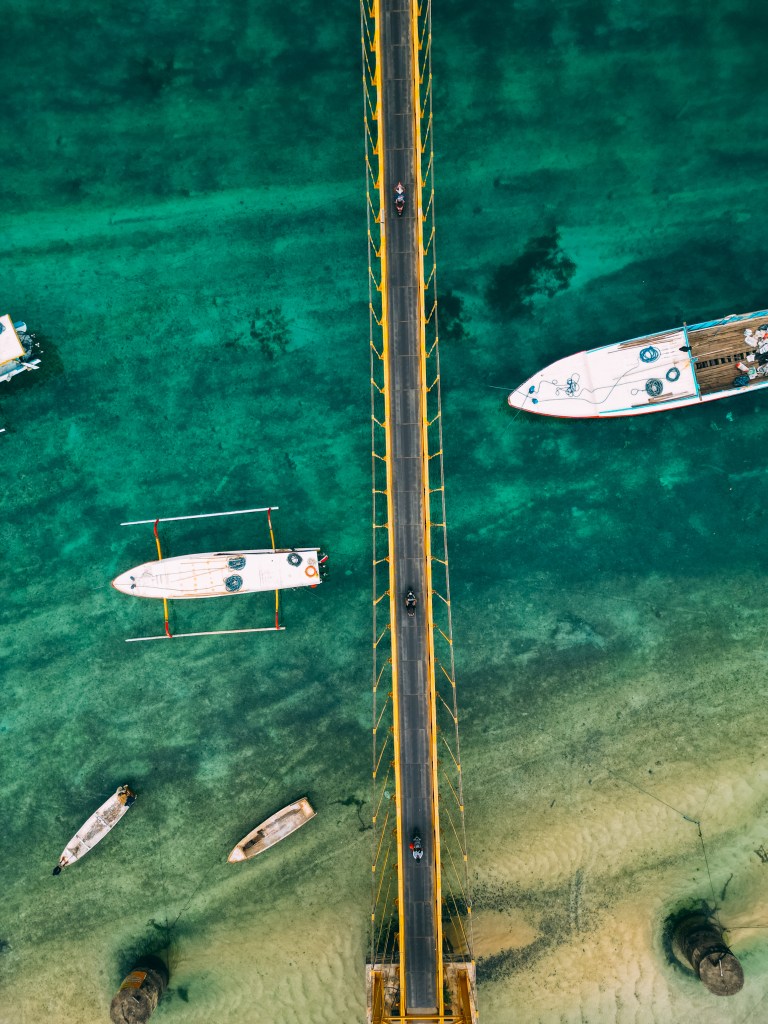 Top-down drone shot of the Yellow Bridge over clear turquoise water with traditional boats beside it, Nusa Ceningan